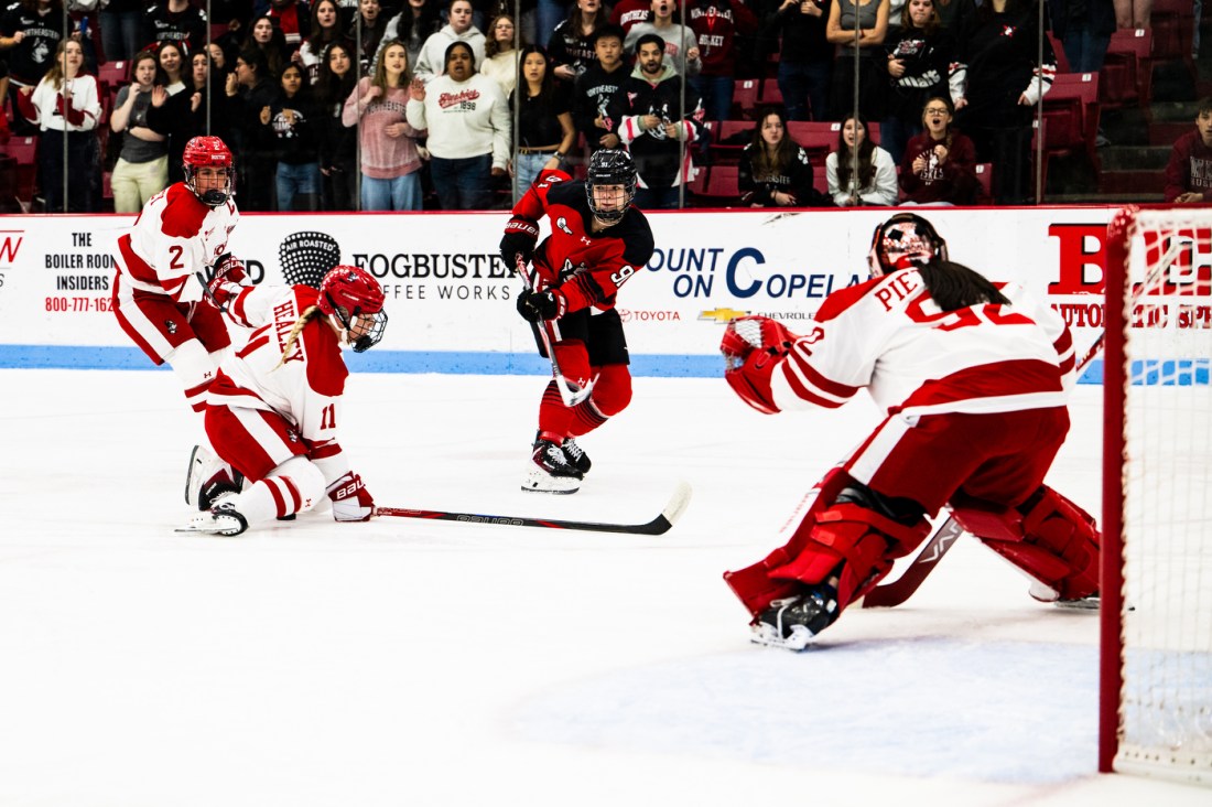 A Northeastern women's hockey player shooting the puck on net against the Boston University goalie. Fans are visible in the background cheering.
