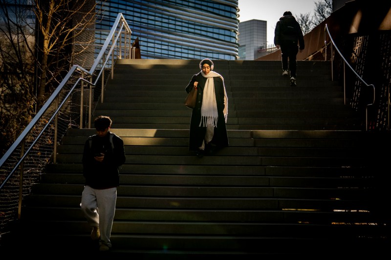A pedestrian walking down stairs on the Boston campus is illuminated by sunlight.