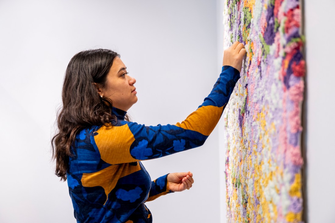 A person adjusts a colorful textile-based data visualization artwork mounted on a gallery wall. 