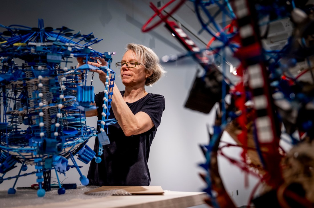 Artist Nathalie Miebach fine-tunes a suspended blue data visualization art sculpture displayed on a table in a gallery space. 