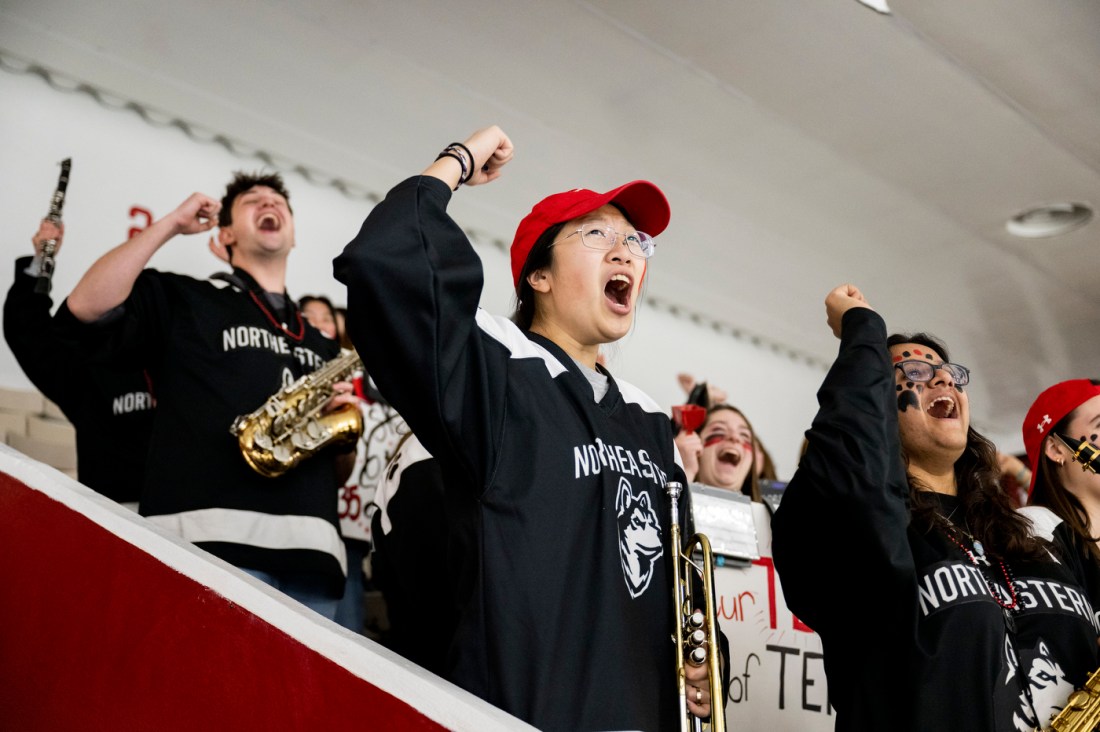 Northeastern hockey fans in the pep band celebrate enthusiastically, wearing black Northeastern shirts and red hats, with one fan playing a saxophone while others raise their arms and cheer.