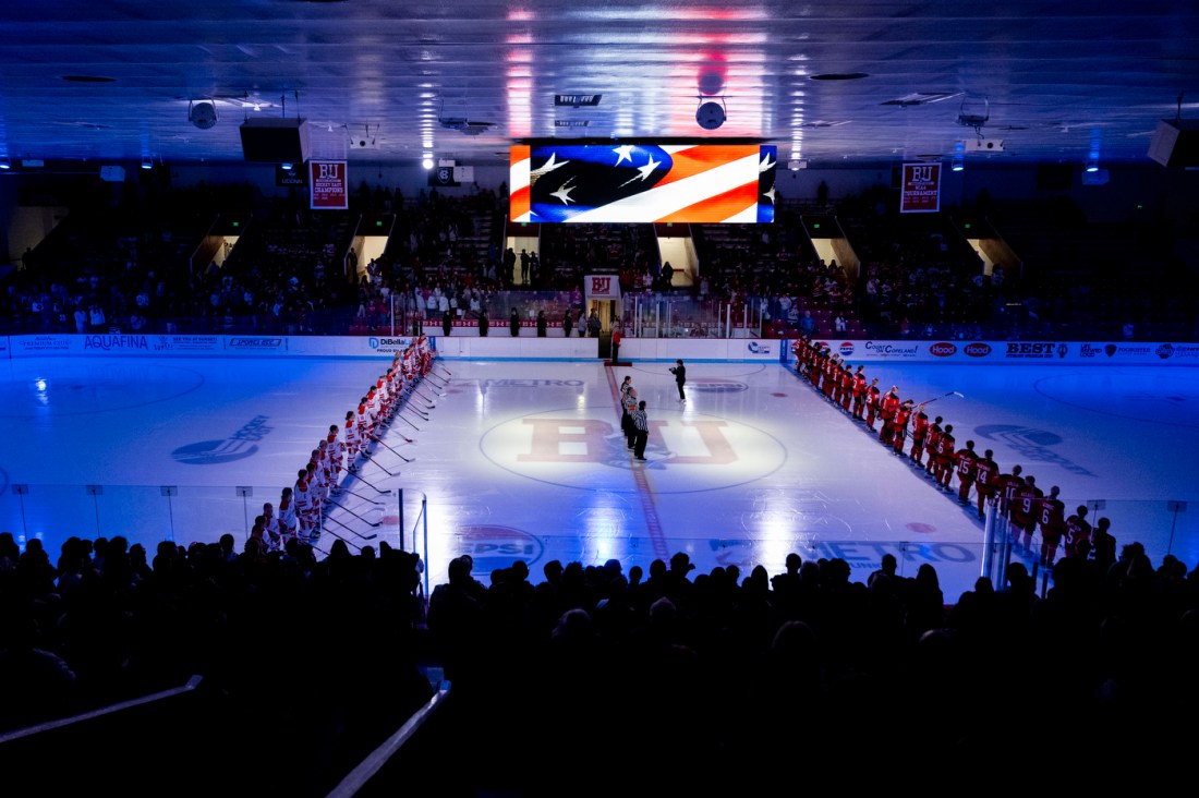 Wide view of the BU hockey arena during the national anthem, showing teams lined up along blue lines on the ice, with dramatic blue lighting, the US flag visible on a screen, and crowd-filled stands.