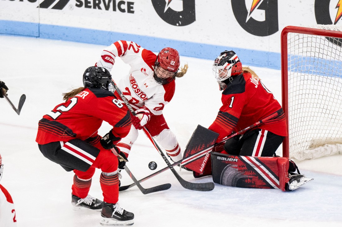 Action shot of a BU women's hockey player battling for the puck near the goal where two Northeastern women's hockey players are defending. 