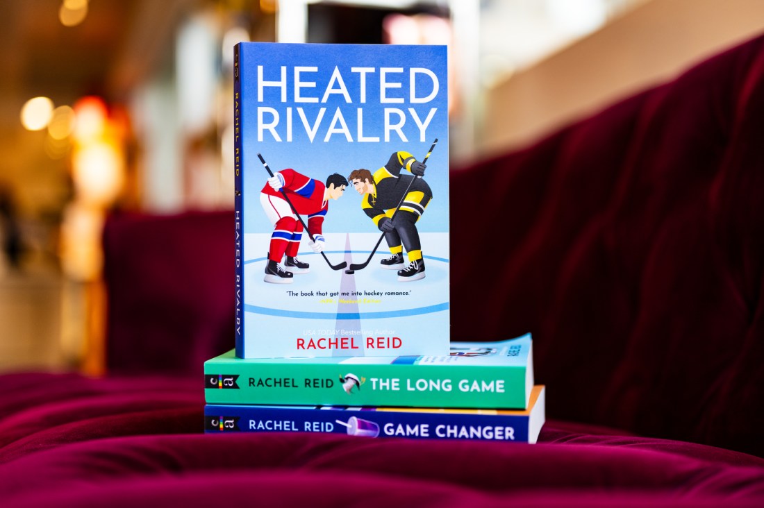 A picture of the heated rivalry book on a stack of other books from the series (The Long Game and Game Changer). The cover features two hockey players in different uniforms facing off.