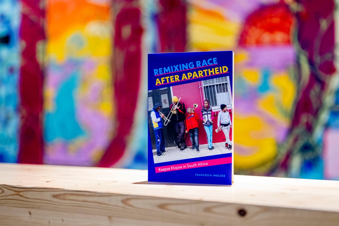 The book "Remixing Race After Apartheid, with a mostly blue cover with a photograph of street musicians, is presented against a very colorful wall.