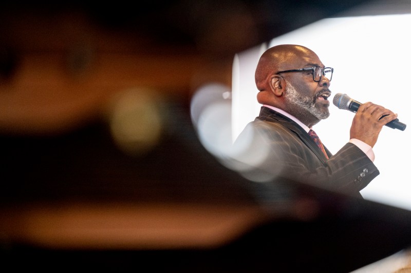 A portrait of a speaker with a microphone addressing an audience.