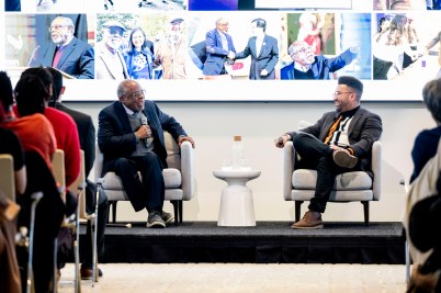 Ted Landsmark (left) and Zolano Kanno-Youngs (right) sitting in two grey armchairs on stage in East Village and laughing. They are both holding microphones.