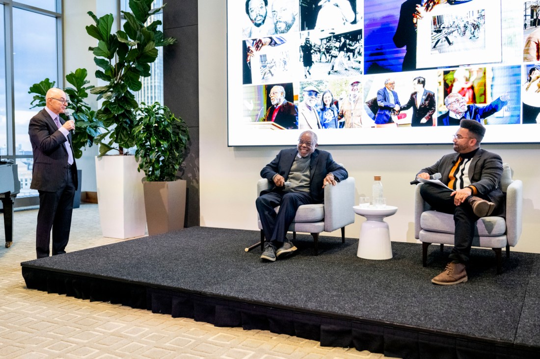 Zolan Kanno-Youngs and Ted Landsmark sitting on stage while President Aoun stands just off stage speaking into a microphone. 