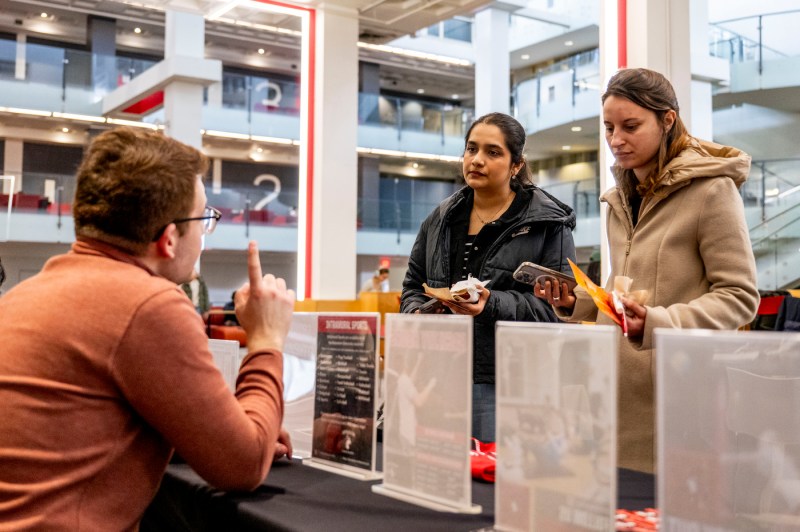 A man talks to two women at a tabling event inside a building.