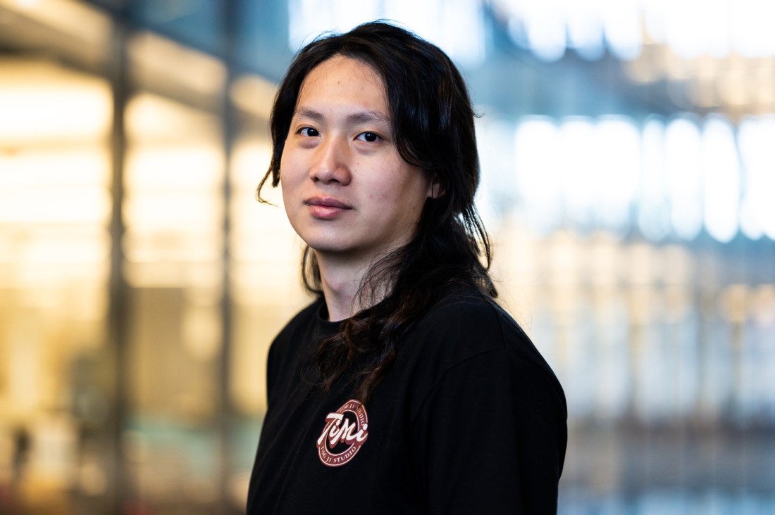 Portrait of a person with long dark hair wearing a black shirt, standing indoors with a softly blurred, glass-and-steel background, looking calmly toward the camera.