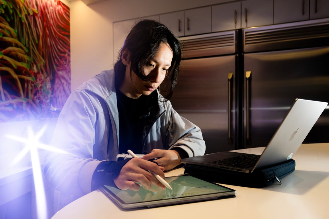 Person with long dark hair sits at a table working on a laptop and writing in a notebook, wearing a light jacket in a modern kitchen space with stainless steel appliances and colorful wall art behind them.