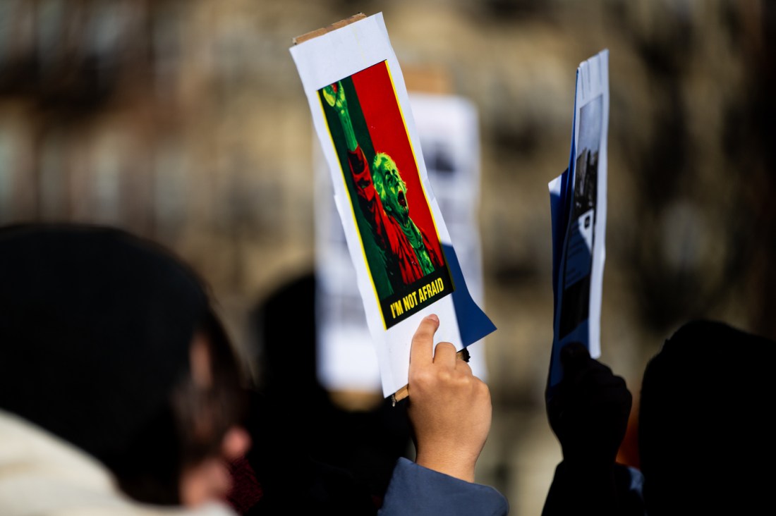 A close-up shows a hand holding a protest sign featuring an image and the words “I Am Not Afraid” at an Iranian student rally.