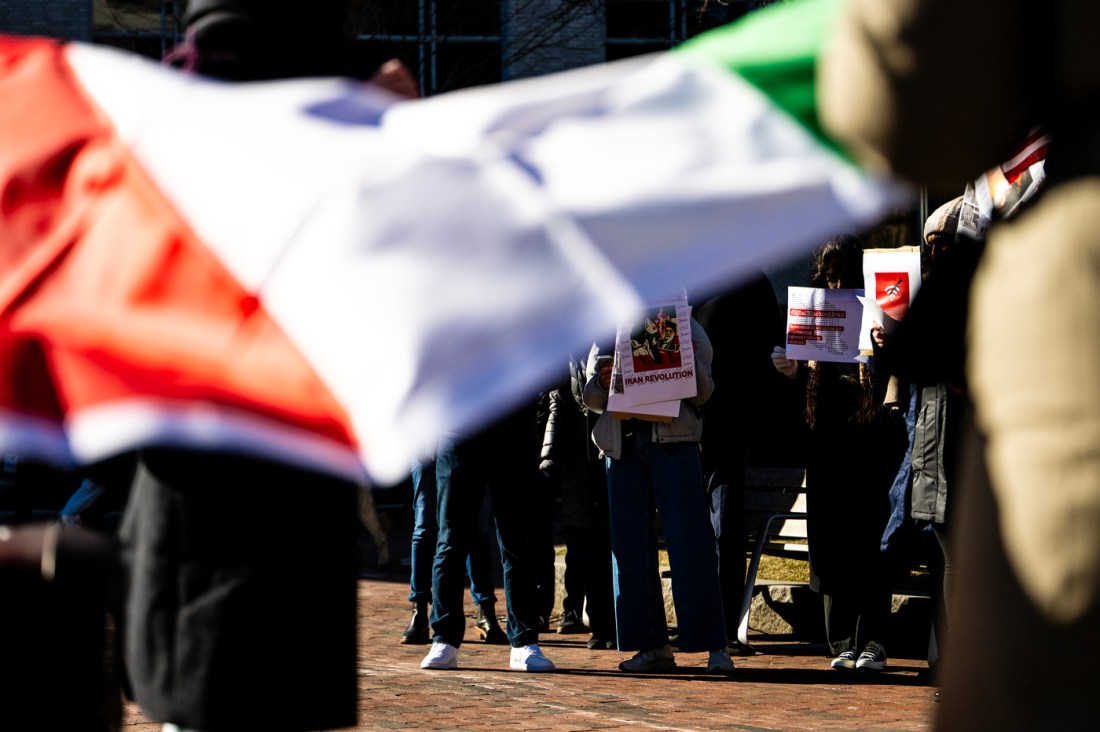 An Iranian flag fills the foreground as students gather holding protest signs in the background at a rally on Krenztman Quad.