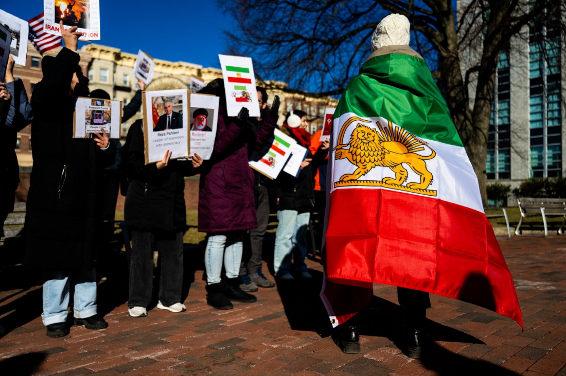 A protester draped in an Iranian flag stands in the foreground as students hold signs during an Iranian student rally on Krenztman Quad.
