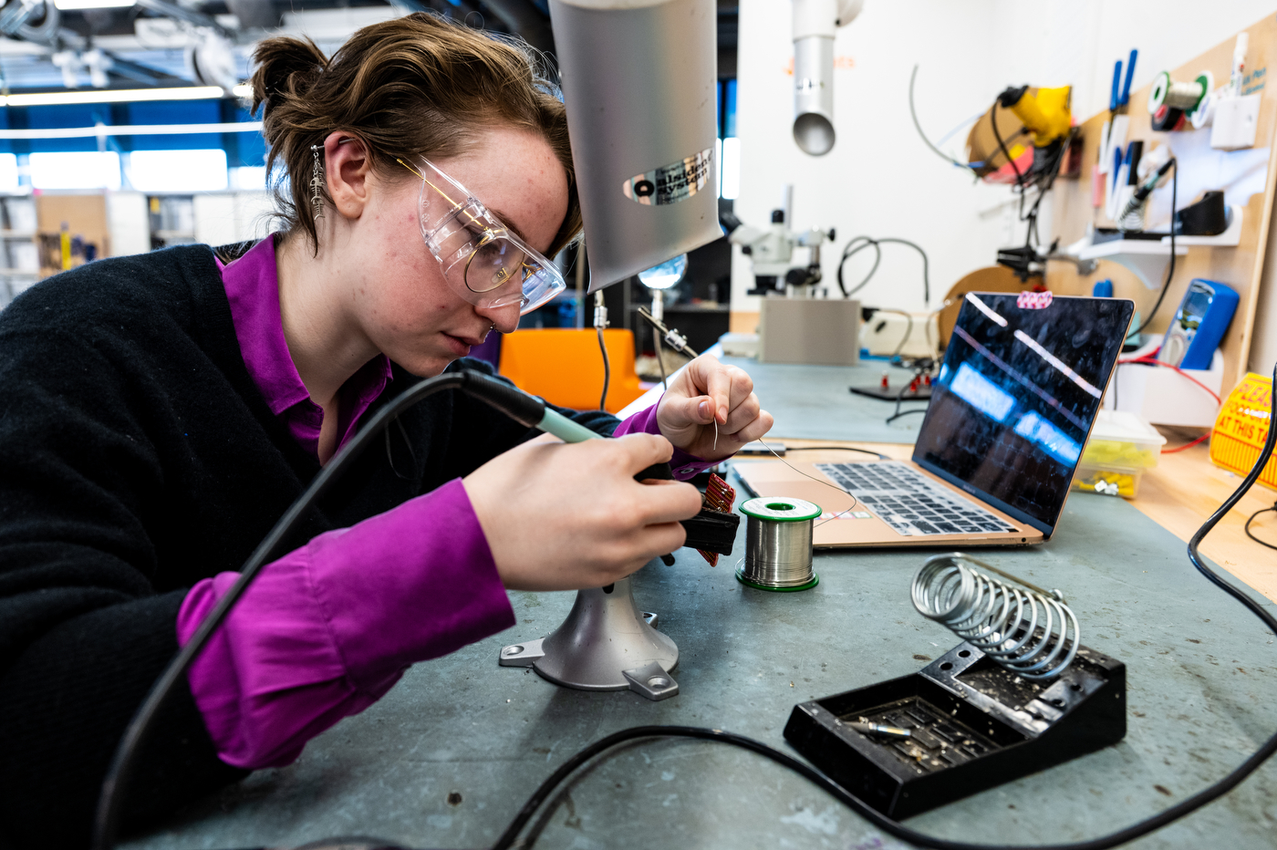 A girl wearing glasses, a black sweater and purple long sleeve shirt sits at a workbench with a laptop open as she is soldering a chip.