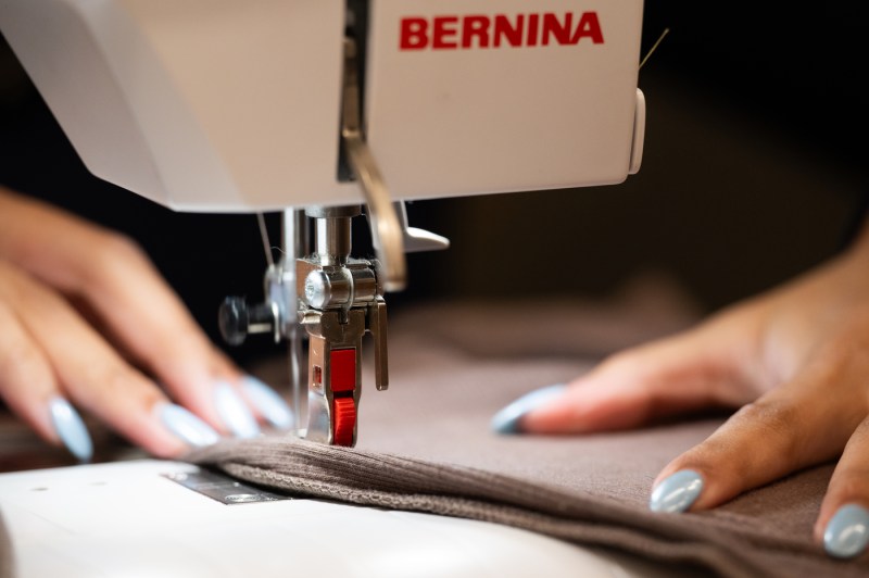 A close up of a sewing machine needle with a brown peice of fabric running through it and a woman's hand with blue nails.
