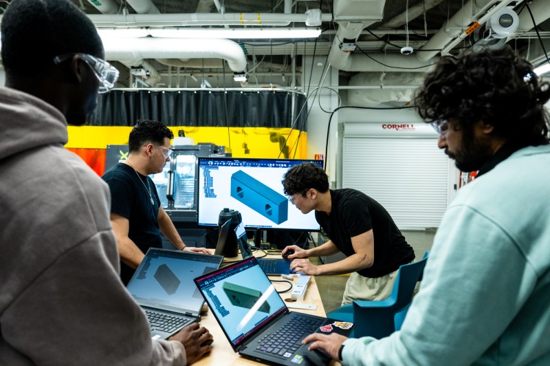 Students stand around laptops and observe a screen with blocks on it during an introduction to welding class.