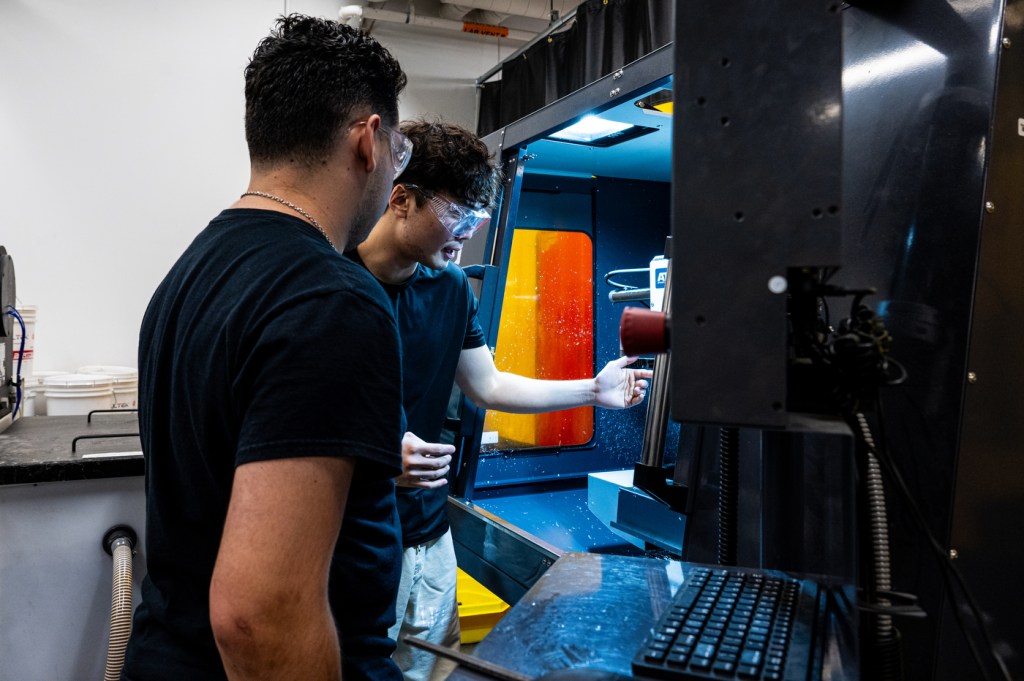 Two men in black T-shirts and in protective glasses look at a piece of equipment.