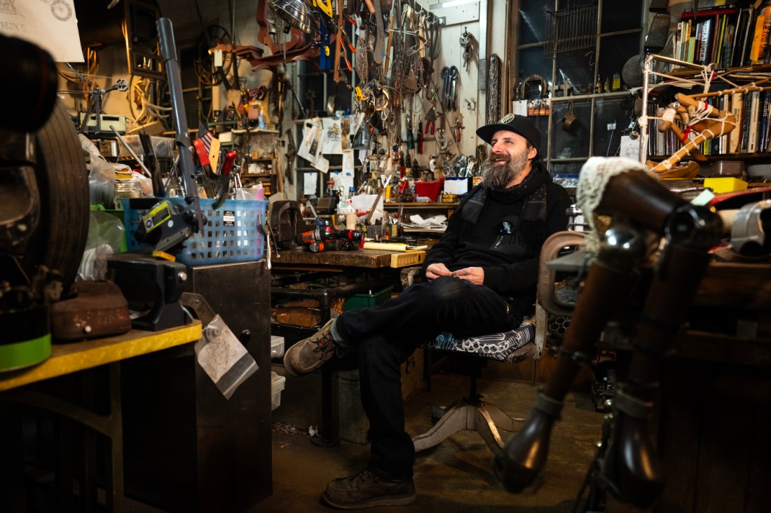 Michael Ulman sits in his studio while smiling and surrounded by metal objects hanging on every surface.