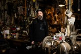 Artist Michael Ulman stands in front of a wall of household objects in his art studio, sporting a long beard, black baseball hat and black hoodie.