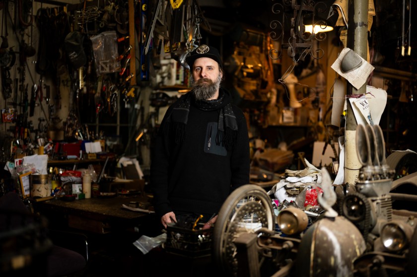 Artist Michael Ulman stands in front of a wall of household objects in his art studio, sporting a long beard, black baseball hat and black hoodie.