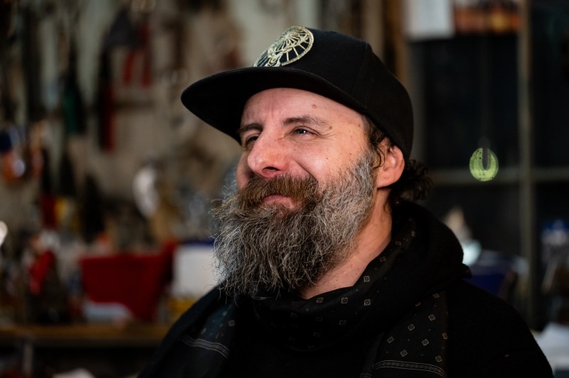 Michael Ulman smiles while wearing a black baseball hat and black hoodie.