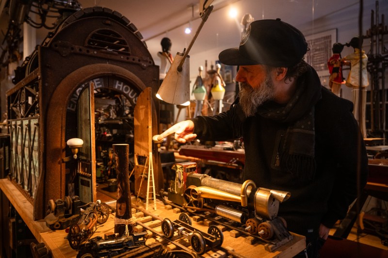 Michael Ulman points at a diorama of a train engine shelter with miniature train tracks coming out of the large double doors.
