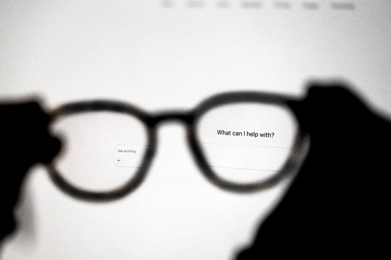 Two silhouetted hands hold a pair of glasses in front of a monitor, with the words 'what can I help with?' in focus on the screen.