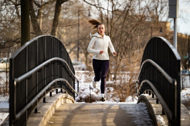 Lily Poteet wearing black leggings, white sneakers and a white zip-up sweater running over a bridge in an urban park.