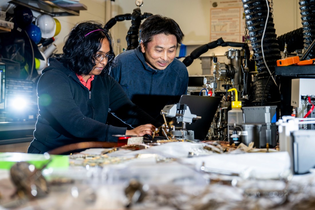 A woman with dark hair works on a computer, next to her is a man in a blue jacket. Both are in a laboratory surrounded by equipment.