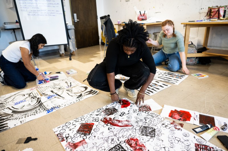 Three students spread paper on the ground, while kneeling down to paint with assorted colors.