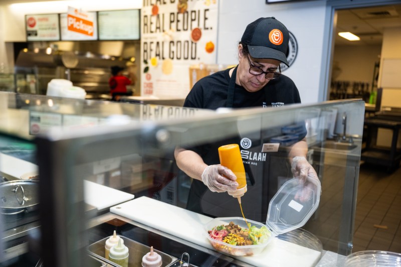 A food service works sprays sauce on a meal in a reusable container.