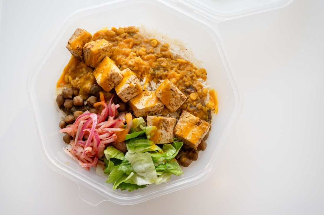 Overhead view of a takeout bowl containing seasoned tofu cubes over white rice, topped with yellow lentil curry sauce and served with lentils, pickled red onions, and fresh lettuce.