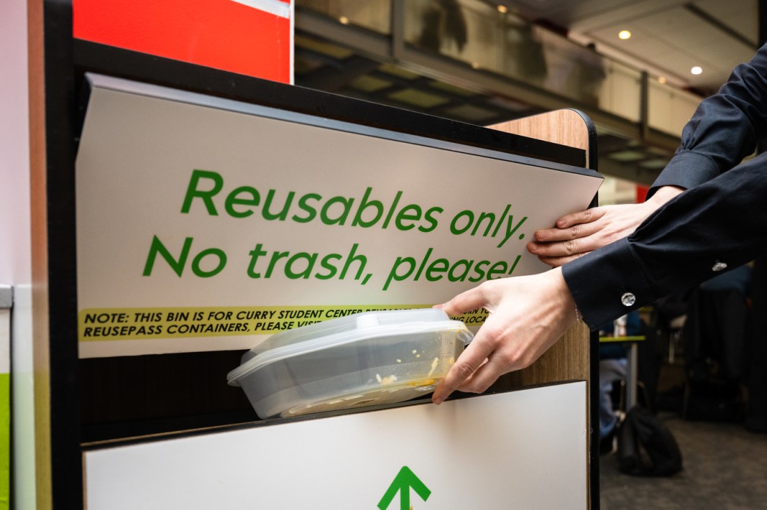 Close-up of hands placing a reusable container into a collection bin marked 'Reusables only. No trash, please!' for the Curry Student Center ReusePass program