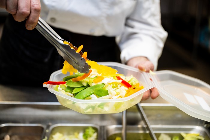 A person uses metal tongs to add grilled chicken strips to a takeout container filled with fresh vegetables including snap peas, bell peppers, and lettuce over white rice.