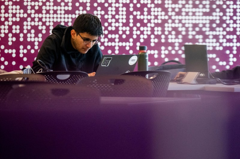 A students sits among purple tables and chairs while leaning over a laptop.