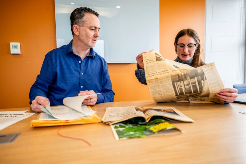 Two people sit at a table examining archival newspapers, with one person holding up a vintage newspaper to view while the other looks through documents, in an office setting with a vibrant orange accent wall and table covered with aged newsprint materials.