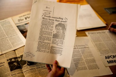 A hand holds up a vintage 1978 newspaper clipping with the headline 'Youth counseling—More harm than good?' featuring a portrait sketch and handwritten note, displayed above a table scattered with other aged newspaper articles.