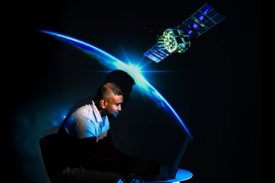 A young man uses his laptop behind a backdrop image of a satellite orbiting Earth.