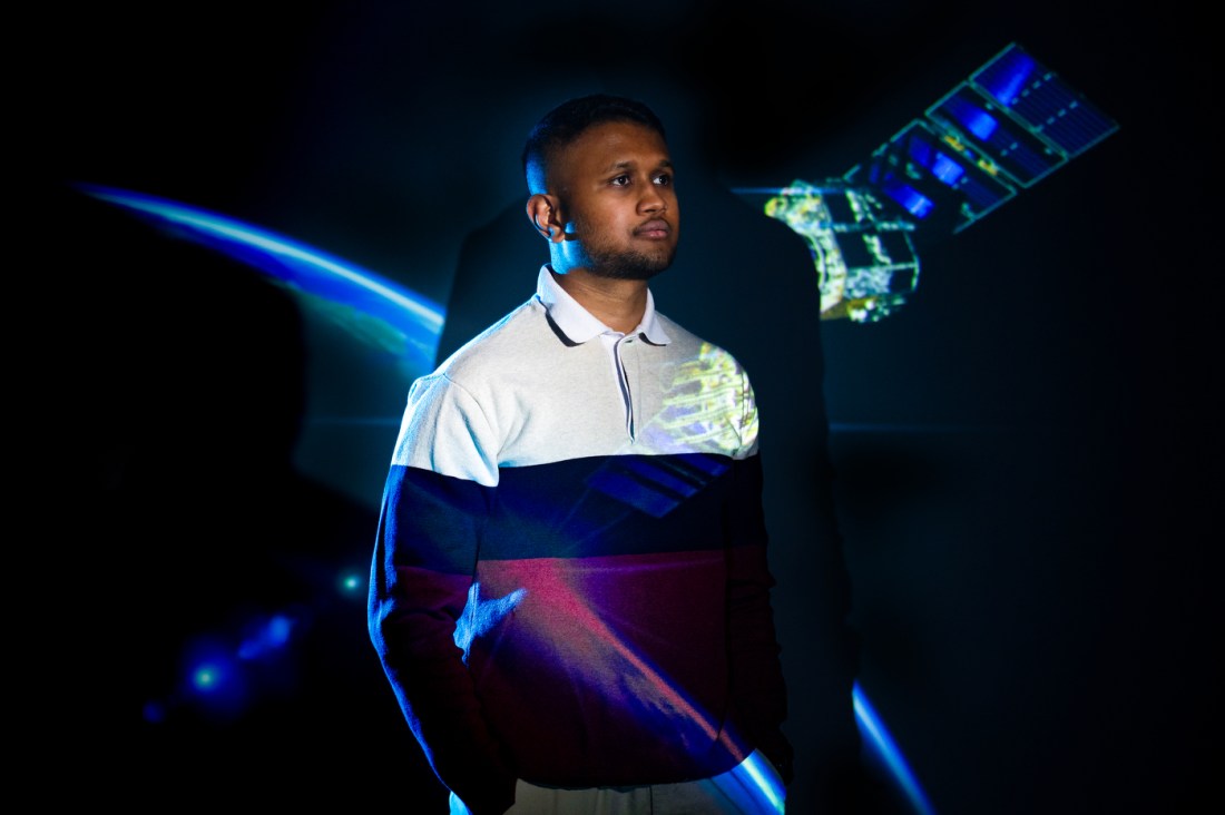A man looks off in the distance with a picture of a satellite orbiting the Earth behind him.  