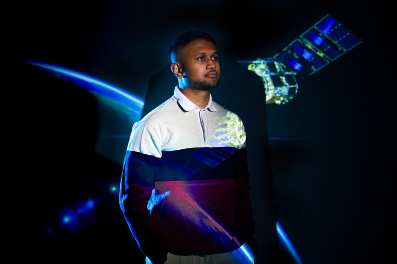 A student poses for a portrait, looking to the right off camera with a projection of the planet and satellite behind him.