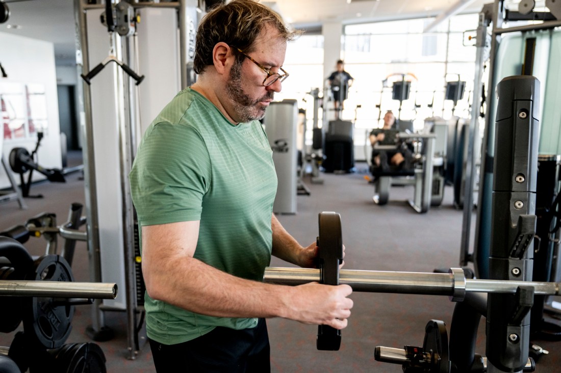 A man in a green t-shirt with glasses positions a weight onto a barbell. 