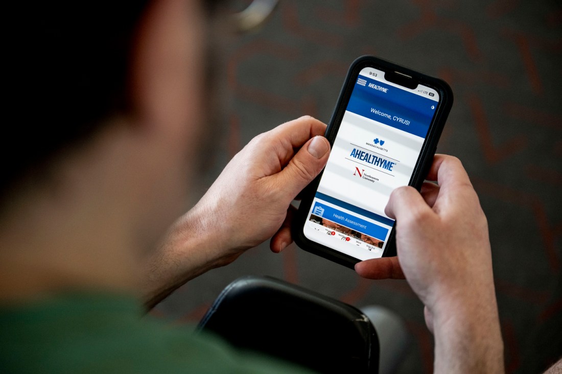 A man's hands holding a smartphone displaying an app that says 'AHealthyMe' on it in all caps blue letters, with the Northeastern logo underneath. 