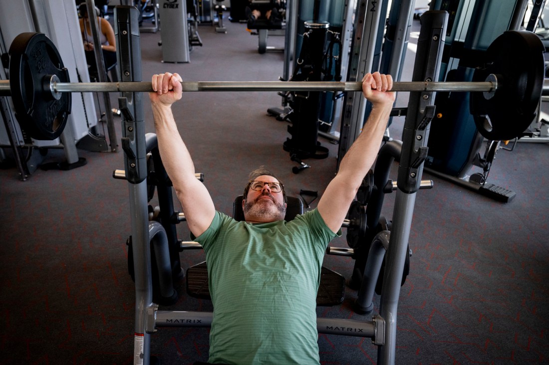 A man performs a bench press exercise on a Smith machine in a gym. He is lying on a flat bench with his arms fully extended overhead, gripping the barbell. He is wearing a light green t-shirt and glasses and he has a beard.