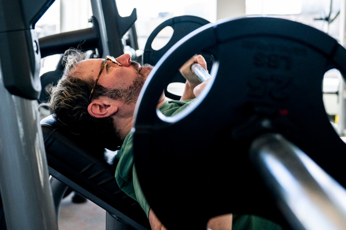 Close-up of a man with glasses and a beard performing bench press with a barbell. 