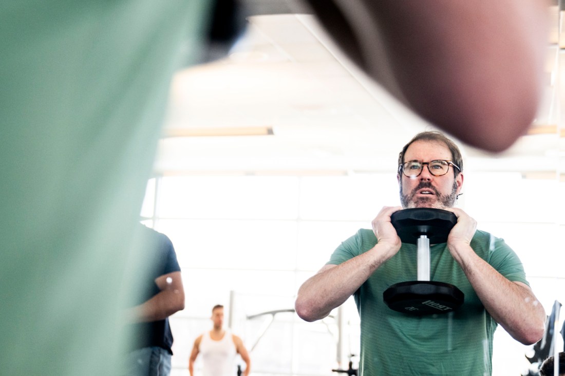 A man in a green t-shirt and glasses holds a dumbbell at chest level with both hands in a gym. 
