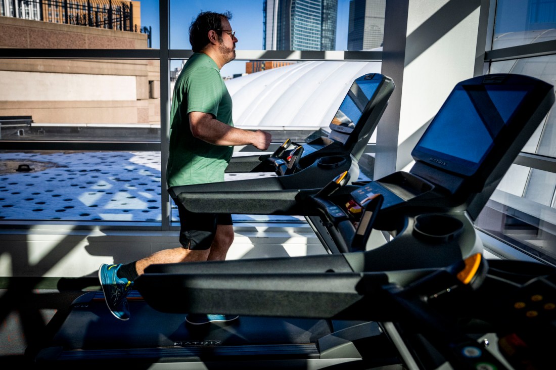 A man in a green shirt and glasses runs on a treatmill at a gym. 