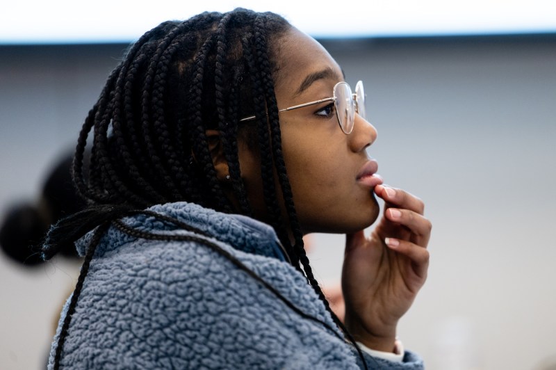 A student with glasses and a sweater touches her chin while listening to the lecture.