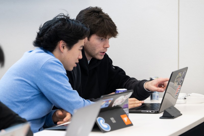 A student in a blue shirt looks at another student's monitor, both discussing something on the screen.