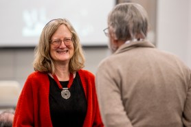 Rabbi Sara Paasche-Orlow, wearing a red cardigan, black top, and silver medal on a red ribbon around her neck, smiling at another person whose back is to the camera. Paasche-Orlow has shoulder-length blonde hair and glasses. 
